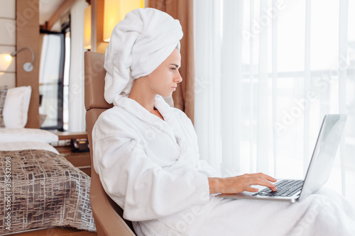 Wise young housewife competently manages house with the help of a personal diary and time management, relaxing after a bath in a bathrobe and a towel with a laptop in her hands. Daily routine concept
