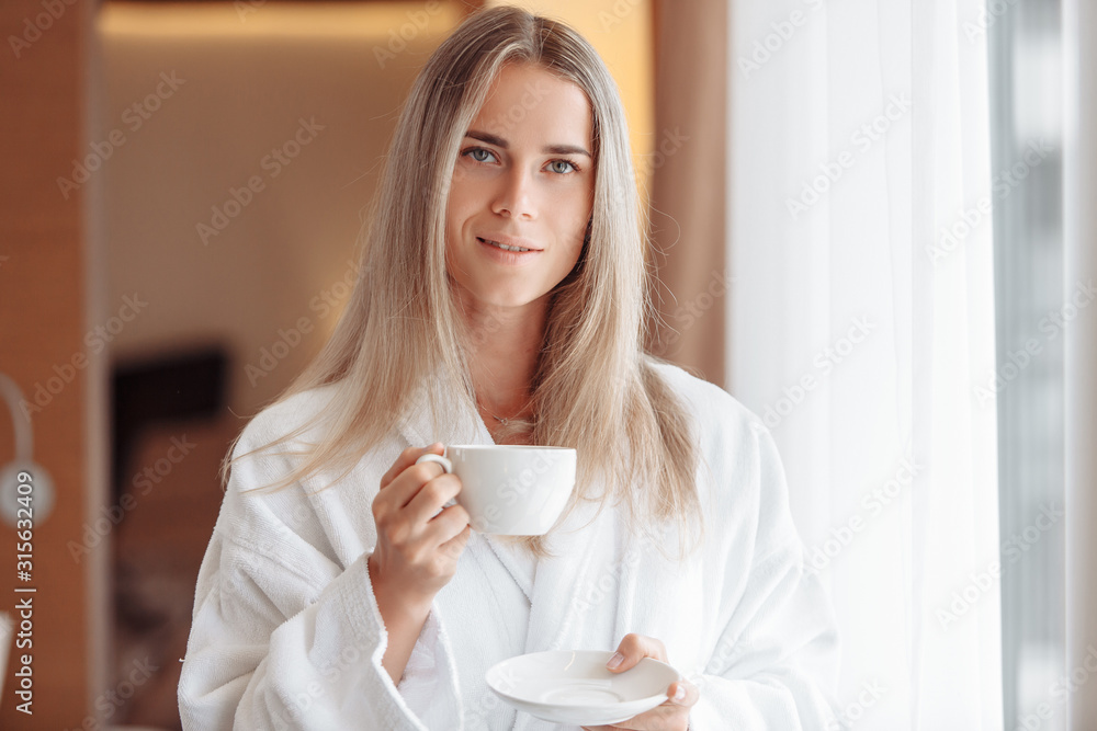 Beautiful young business girl is drinking coffee in morning. Charming woman in good mood starts day with a smile, looks out bedroom window. An invigorating drink helps set up for work Beautiful young business girl is drinking coffee in morning. Charming woman in good mood starts day with a smile, looks out bedroom window. An invigorating drink helps set up for work