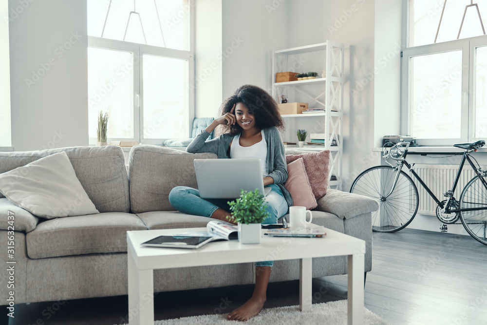 Beautiful young African woman in casual clothing using laptop while resting at home Beautiful young African woman in casual clothing using laptop while resting at home