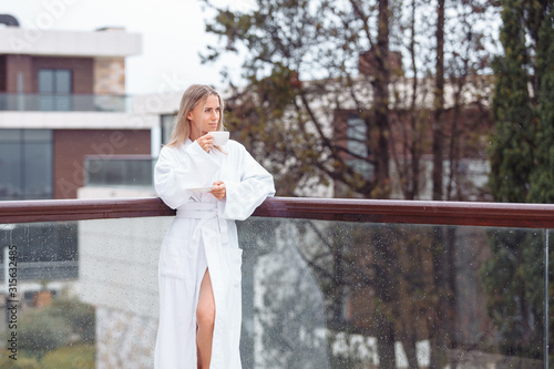 Satisfied beautiful young caucasian woman in coat drinking medicinal tea enjoying beautiful view from hotel spa terrace in morning. Concept of therapeutic rest getting rid of chronic diseases, fatigue © Rithor