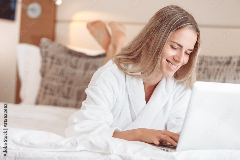Cheerful beautiful young woman freelancer in white coat lies on bed and looks at orders and reviews using laptop and high-speed internet. Concept of personal affairs and independent earnings. Cheerful beautiful young woman freelancer in white coat lies on bed and looks at orders and reviews using laptop and high-speed internet. Concept of personal affairs and independent earnings.