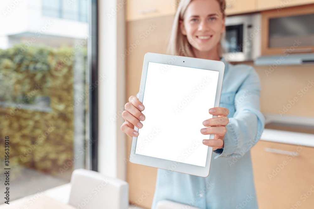 Young happy blurred caucasian girl holding a tablet with a white screen while standing in her kitchen demonstrating a smart home system that saves time. Smart home system concept Young happy blurred caucasian girl holding a tablet with a white screen while standing in her kitchen demonstrating a smart home system that saves time. Smart home system concept