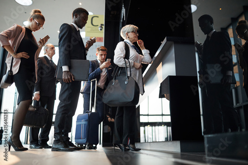 Senior woman with luggage standing near the reception and registering with other people standing in a queue at the airport