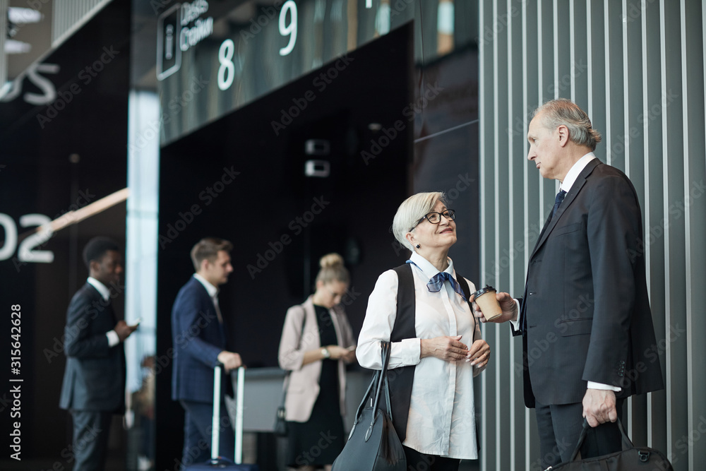 Senior businessman talking to senior businesswoman and drinking coffee while they waiting for the registration at the airport