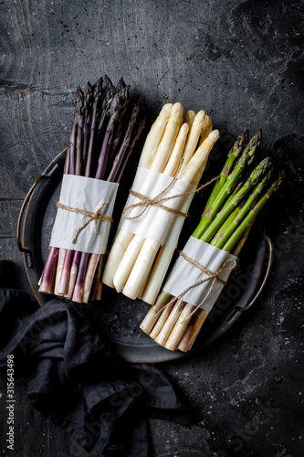 Bunches of fresh green, purple, white asparagus on vintage metal tray over dark grey rustic background. Top view, copy space