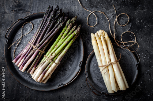 Bunches of fresh green, purple, white asparagus on vintage metal tray over dark grey rustic background. Top view, copy space