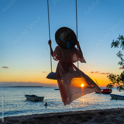 Beautiful girl in a straw hat and pareo swinging on a swing on the beach during sunset of Zanzibar island, Tanzania, Africa. Travel and vacation concept