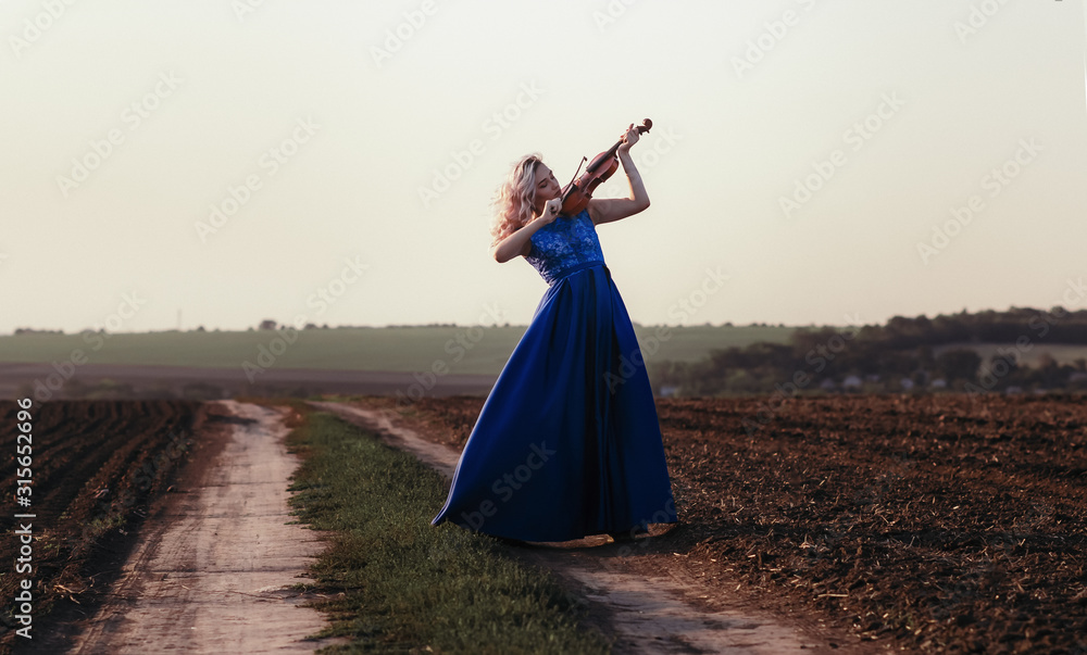 Fototapeta premium woman in long dress playing violin on background of field path with a club of dust, girl engaged in musical art, performance on nature, concept passion in music