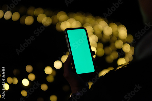over shoulder of one hand holding green screen phone in darkness. Golden bokeh background