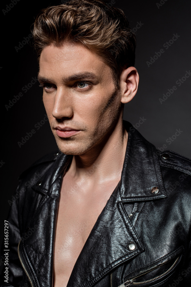 sexy young man in biker jacket looking away on black background