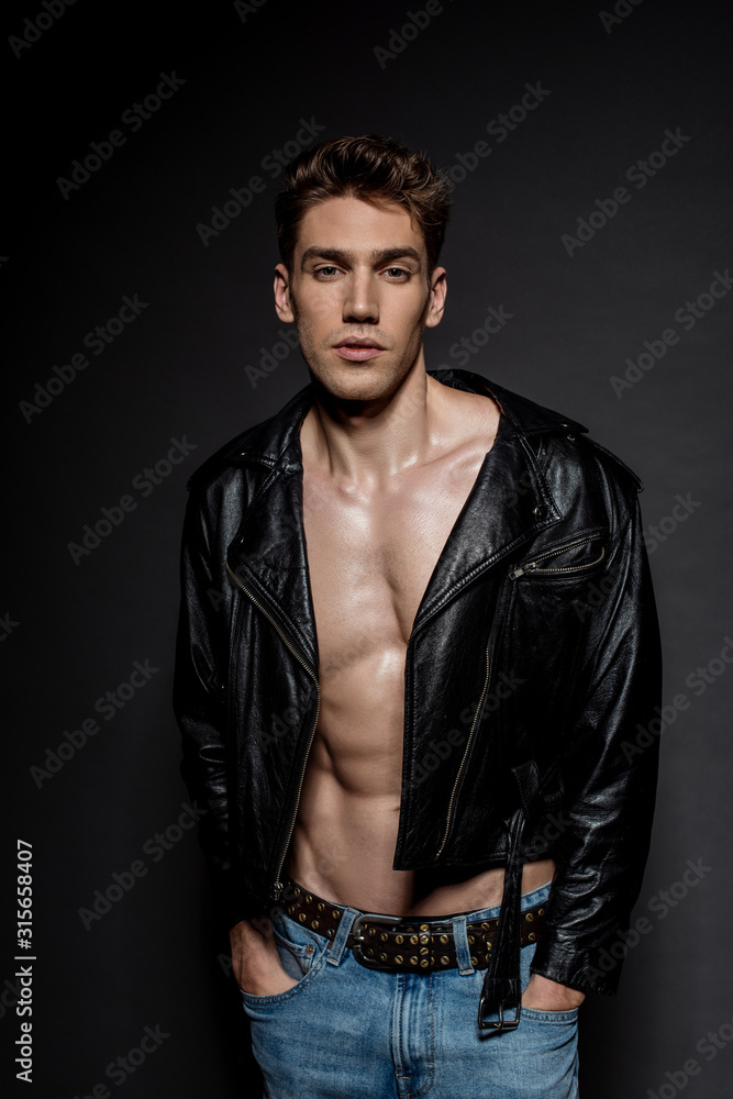 sexy young man with muscular torso in biker jacket and jeans posing with hands in pockets on black background