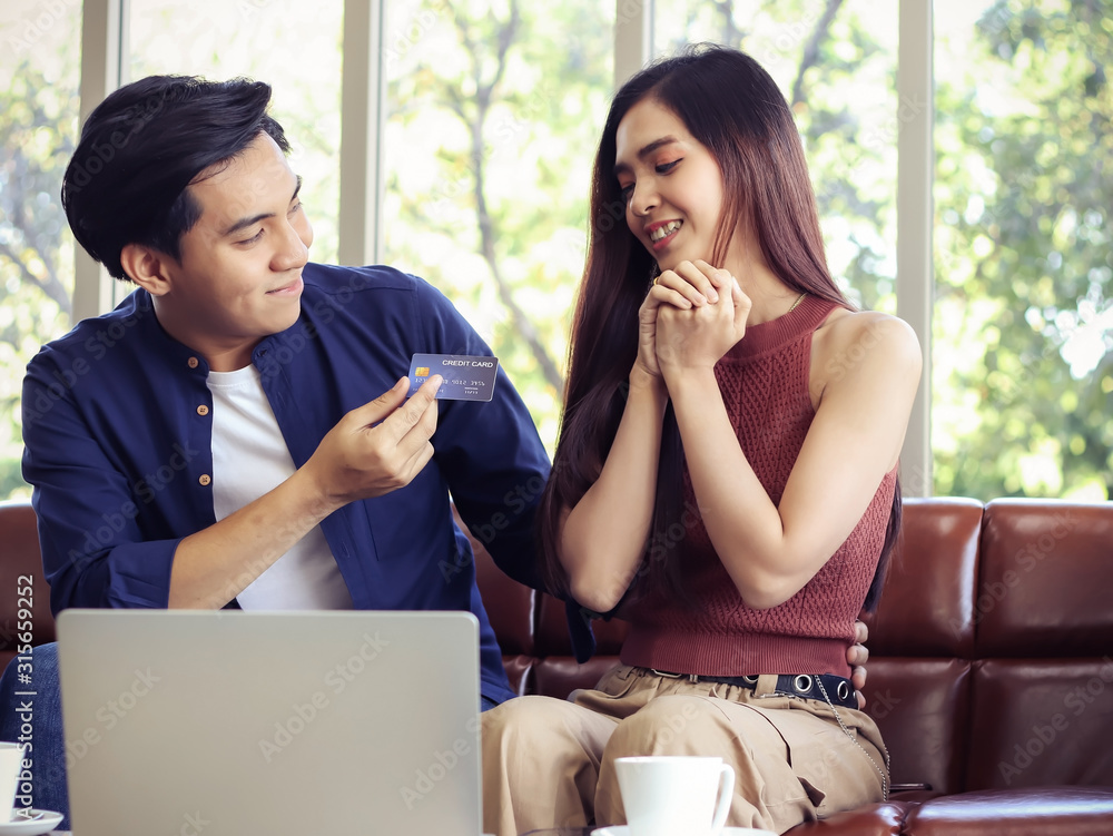 ttractive couple sitting close together on couch with computer laptop on the table , a man giving credit card to his girlfriend for shopping making her feel glad. ttractive couple sitting close together on couch with computer laptop on the table , a man giving credit card to his girlfriend for shopping making her feel glad.