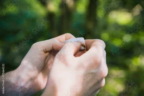 Man under the influence of drugs or alcohol struggling to roll a tobacco cigarette or marijuana joint or spliff in the wood forest with nice sunny green bokeh background pale young hands