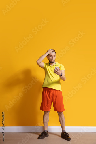 Shocked, wondered. Young caucasian man using smartphone, serfing, chatting, betting. Full length portrait isolated on yellow background. Concept of modern technologies, millennials, social media.
