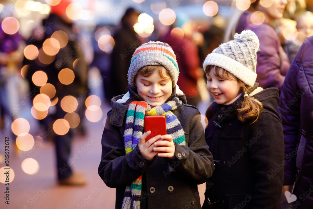 Two little kids, boy and girl having fun on traditional Christmas market during strong snowfall with taking photos with mobile smartphone or phone. Happy children, siblings and best friends Two little kids, boy and girl having fun on traditional Christmas market during strong snowfall with taking photos with mobile smartphone or phone. Happy children, siblings and best friends