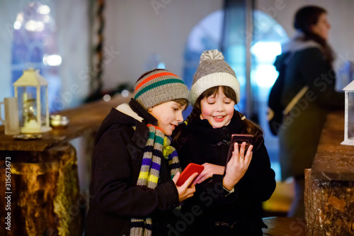 Two little kids, boy and girl having fun on traditional Christmas market during strong snowfall with taking photos with mobile smartphone or phone. Happy children, siblings and best friends