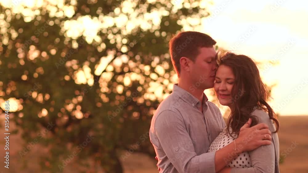 Portrait of a happy pregnant couple in a field at sunset, with a blurred tree in the background. Red light. A pregnant woman with her beloved husband enjoys a walk outside the city at sunset. Portrait of a happy pregnant couple in a field at sunset, with a blurred tree in the background. Red light. A pregnant woman with her beloved husband enjoys a walk outside the city at sunset.