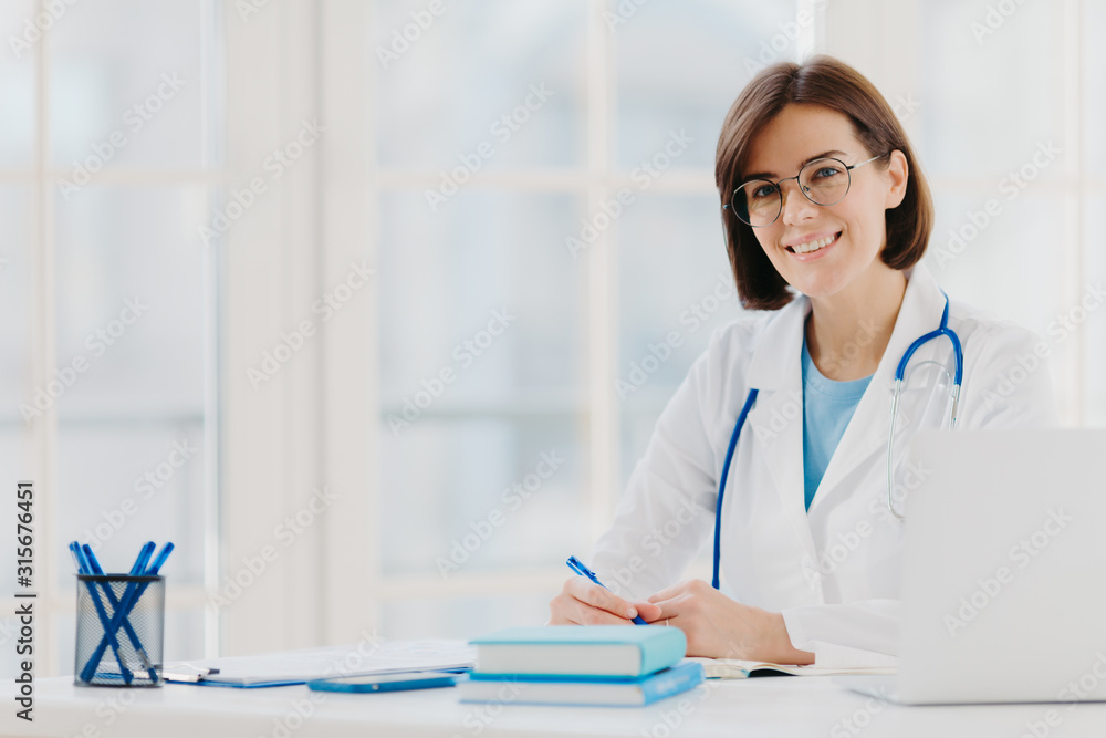 Professional woman doctor writes down notes, poses at desktop in office with laptop, wears white coat, spectacles and phonendoscope around neck, looks through medical documents. Healthcare concept Professional woman doctor writes down notes, poses at desktop in office with laptop, wears white coat, spectacles and phonendoscope around neck, looks through medical documents. Healthcare concept
