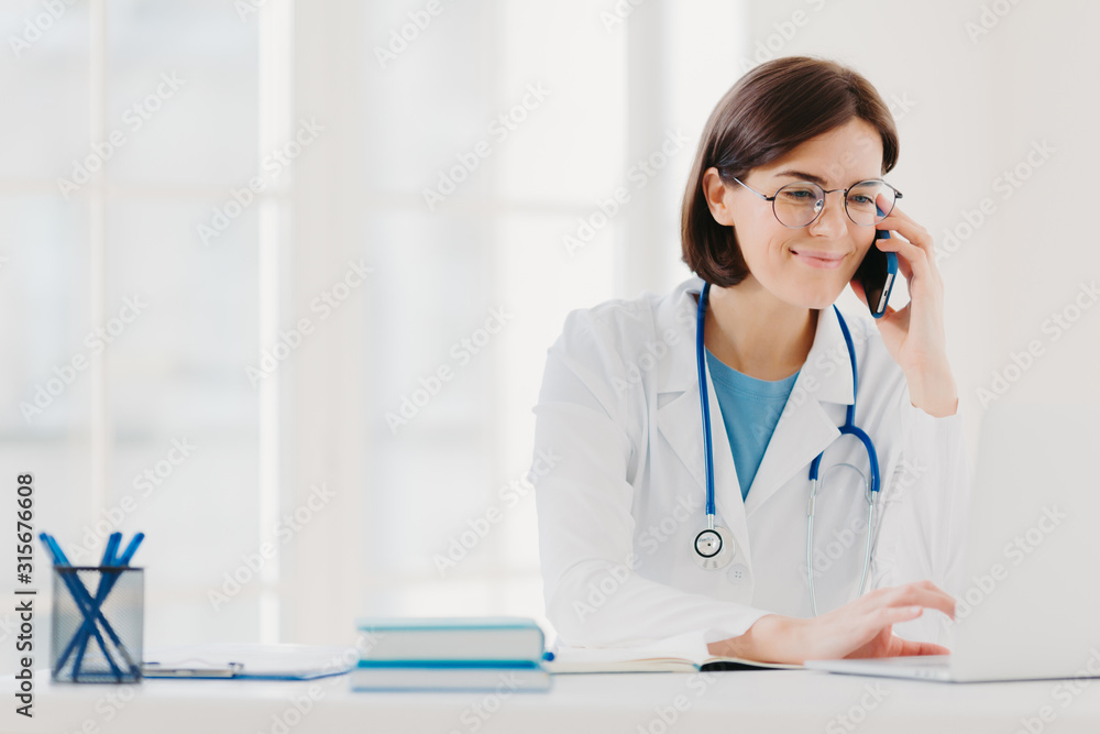 Smiling therapist focused at screen of laptop computer, calls via modern smartphone, wears medical gown, transparent glasses, sits at coworking space, surfes internet. Copy space for your information Smiling therapist focused at screen of laptop computer, calls via modern smartphone, wears medical gown, transparent glasses, sits at coworking space, surfes internet. Copy space for your information