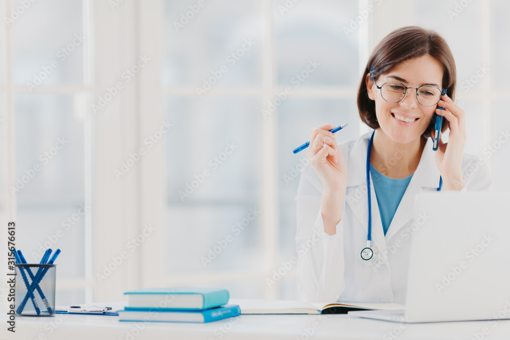 Photo of glad woman therapist or physician has phone conversation with patient, gives advice how to cure disease, wears white gown, reads information on laptop computer, works in private clinic Photo of glad woman therapist or physician has phone conversation with patient, gives advice how to cure disease, wears white gown, reads information on laptop computer, works in private clinic