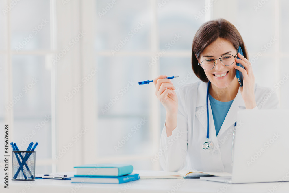 Glad professional doctor concentrated in modern laptop computer, reads useful information, has telephone conversation, discusses medical issues, sits at hospital office with notepads on table Glad professional doctor concentrated in modern laptop computer, reads useful information, has telephone conversation, discusses medical issues, sits at hospital office with notepads on table