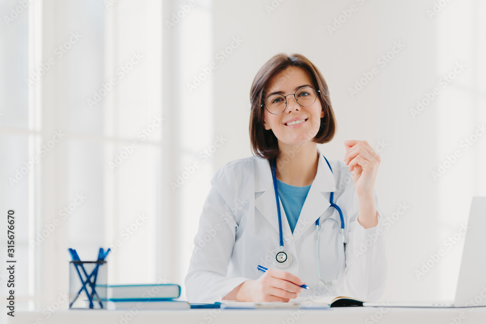 Young woman doctor writes on paper in clinic, creats prescription for patient, does paperwork in hospital, wears medical white gown, sits at cozy modern office. People, occupation, job concept Young woman doctor writes on paper in clinic, creats prescription for patient, does paperwork in hospital, wears medical white gown, sits at cozy modern office. People, occupation, job concept