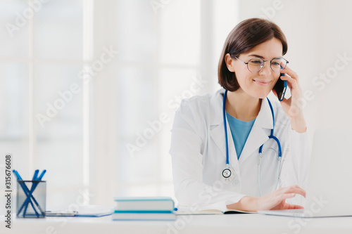 Smiling therapist focused at screen of laptop computer, calls via modern smartphone, wears medical gown, transparent glasses, sits at coworking space, surfes internet. Copy space for your information