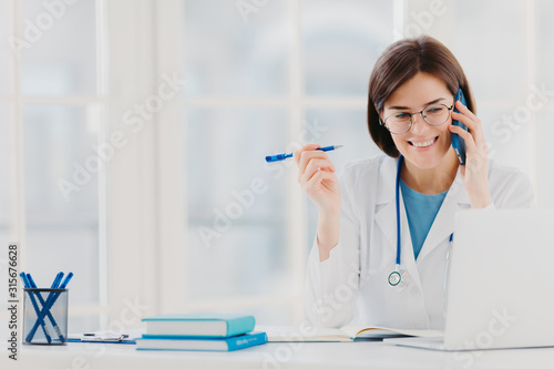 Glad professional doctor concentrated in modern laptop computer  reads useful information  has telephone conversation  discusses medical issues  sits at hospital office with notepads on table