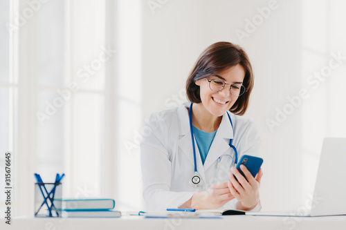Positive female medical worker involved in chatting  holds mobile phone and sends messages  wears medical gown  has friendly cheerful expression  poses at desktop gives online consultation for patient