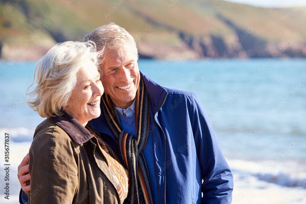 Loving Senior Couple Hugging As They Walk Along Shoreline Of Beach By Waves Loving Senior Couple Hugging As They Walk Along Shoreline Of Beach By Waves