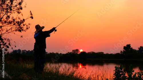 Silhouette fisherman with hat fishing on river bank and blurred sunset sky background in evening time