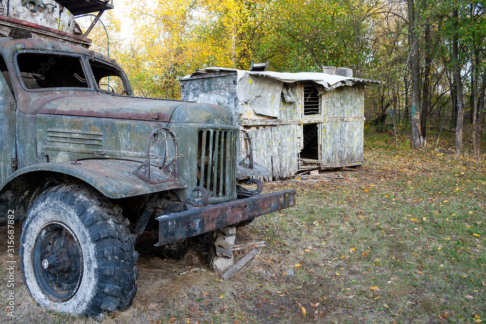 The body of an old dirty truck and a rickety wooden shed of old boards with holes in the walls and with spots of yellow paint is standing on the grass in the forest. Shelter at the tactical paintball