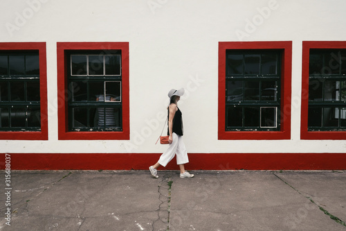 Young elegance woman wearing white hat and trousers walking near city wall with red windows