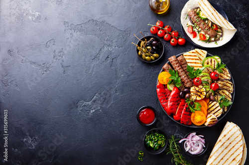 Meat kebab, vegetables on a black plate with tortillas, flat bread. Slate stone background. Copy space. Top view.