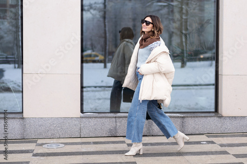 European model girl in a beige oversized down jacket, knitted sweater, flared jeans with a handbag and glasses is walking down the street. Life style