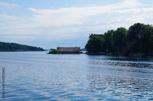 A view on the Chiemsee lake (Bavaria, Germany). In the distance, a cottage can be seen, as well as forested islands. The sky is very blue, there are scattered clouds. Clam waters with slight ripples.