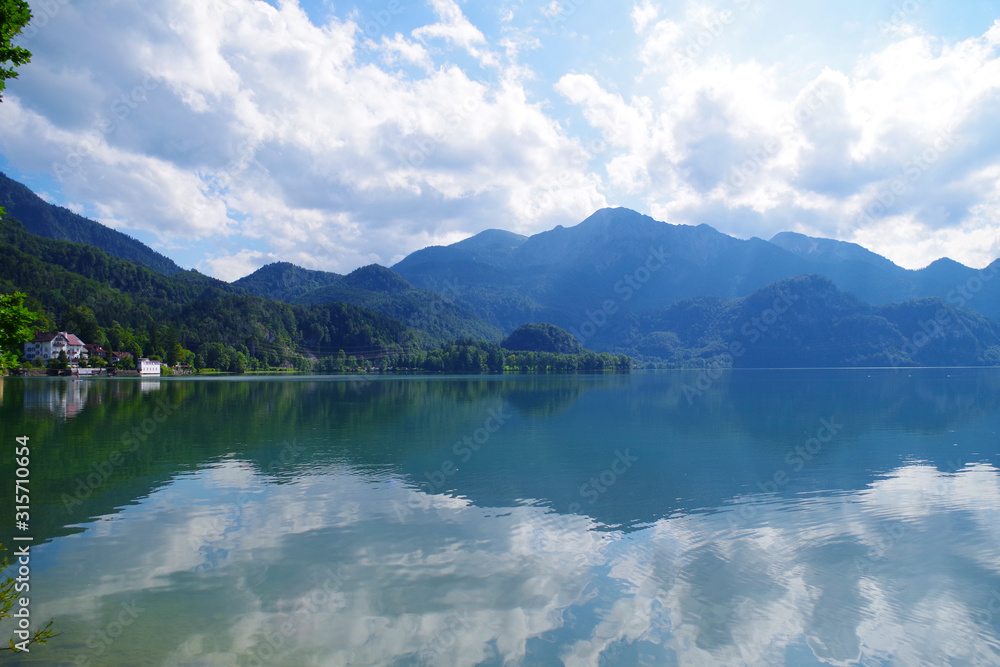 A landscape with a beautiful blue lake surrounded by forested green mountains. In the distance, several  buildings hidden between the trees. It's a bright summer day with some clouds in the sky.