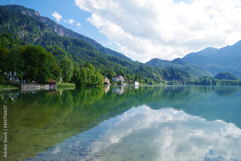 A landscape with a beautiful blue lake surrounded by forested green mountains. In the distance, several  buildings hidden between the trees. It's a bright summer day with some clouds in the sky.