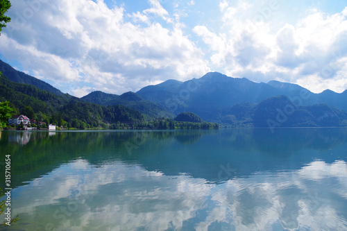A landscape with a beautiful blue lake surrounded by forested green mountains. In the distance, several buildings hidden between the trees. It's a bright summer day with some clouds in the sky.
