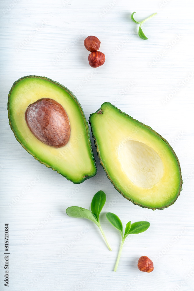 Ripe avocado cut into two parts on a white wooden background with nuts and leaves, one part with a bone.