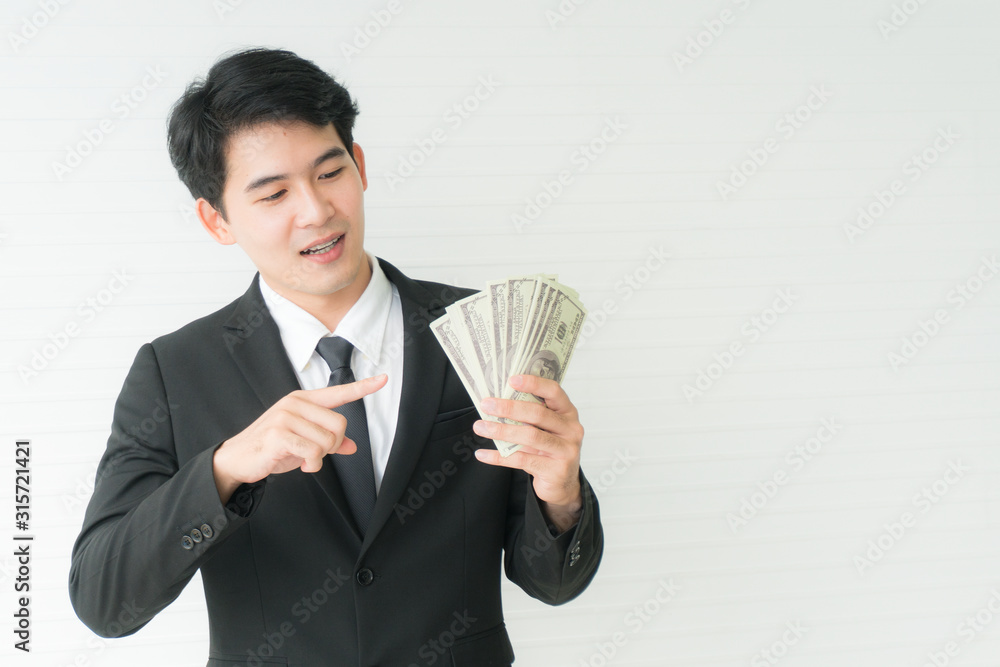 Asian young businessman standing and smiling with point one fingers to dollars one hand holding money dollars .Portrait on white background with copy space for text and advertisement Asian young businessman standing and smiling with point one fingers to dollars one hand holding money dollars .Portrait on white background with copy space for text and advertisement