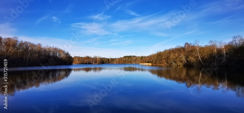 A quiet calm lake in January with different dark and light blue tones sky and trees that are reflected on the shore in the water and magnificent panorama with far-sightedness