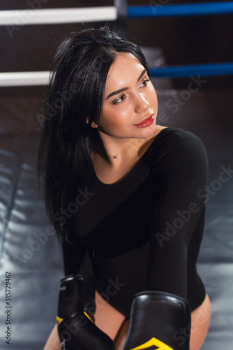 Sexy fighter girl in a black bodysuit and boxing gloves posing in a dark black boxing ring. Long hair woman fitness model. The concept of a healthy lifestyle