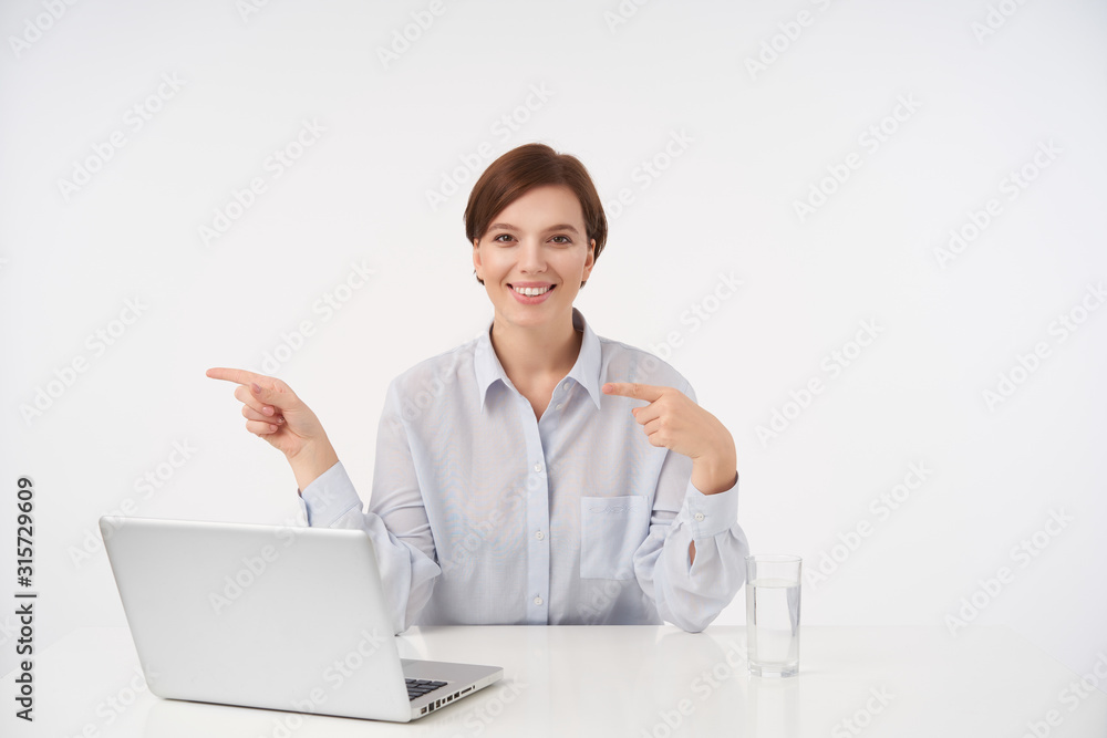 Cheerful young pretty short haired brunette woman working in office with her laptop and looking positively at camera with wide smile, poiting aside with forefingers while sitting over white background
