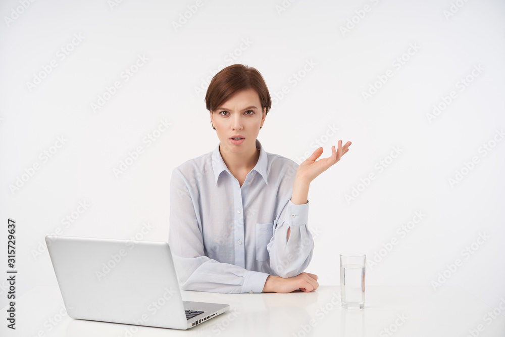 Serious young pretty brown haired female with short trendy haircut frowning scowlingly her eyebrows and keeping palm raised while sitting at table over white background Serious young pretty brown haired female with short trendy haircut frowning scowlingly her eyebrows and keeping palm raised while sitting at table over white background