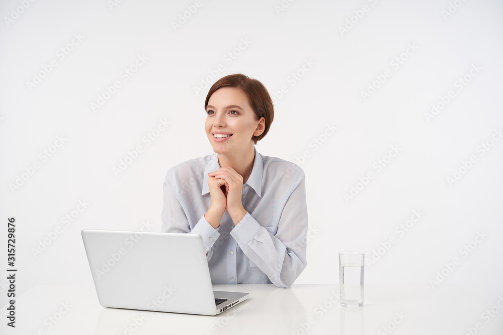 Indoor photo of charming cheerful young brunette female with short trendy haircut folding raised hands and looking aside with pleasant smile, dressed in formal over white background Indoor photo of charming cheerful young brunette female with short trendy haircut folding raised hands and looking aside with pleasant smile, dressed in formal over white background