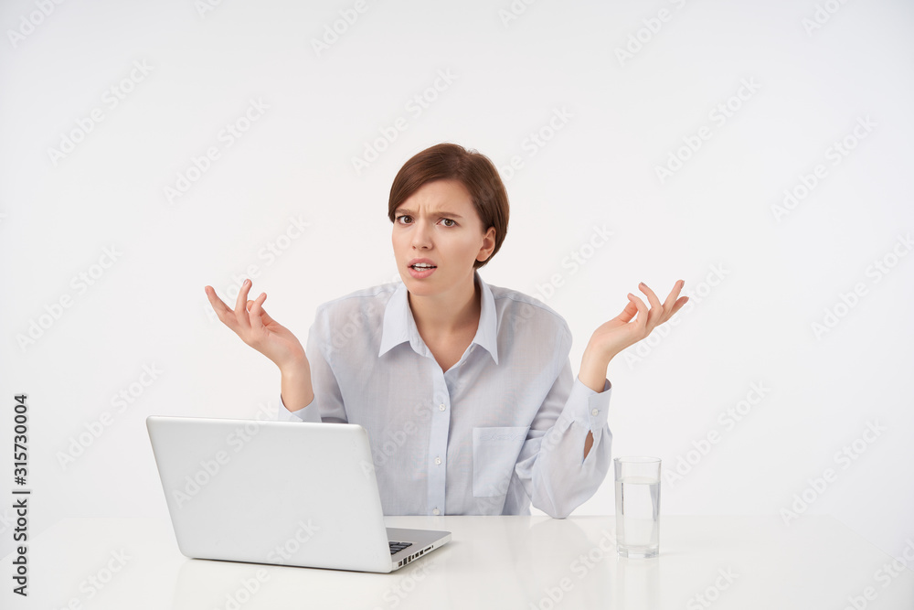 Puzzled young brown-eyed short haired woman with natural makeup looking at camera with displeased face and raising confusedly palms while sitting over white background Puzzled young brown-eyed short haired woman with natural makeup looking at camera with displeased face and raising confusedly palms while sitting over white background