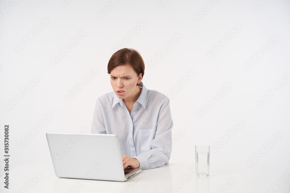 Shot of confused young short haired brunette lady with casual hairstyle looking at screen of her laptop with displeased face, reading unpleasant news, isolated over white background Shot of confused young short haired brunette lady with casual hairstyle looking at screen of her laptop with displeased face, reading unpleasant news, isolated over white background