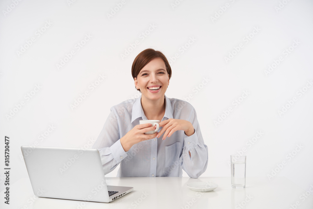 Joyful young attractive brown haired woman with casual hairstyle laughing happily while drinking coffee during working day, dressed in blue shirt while posing over white background Joyful young attractive brown haired woman with casual hairstyle laughing happily while drinking coffee during working day, dressed in blue shirt while posing over white background