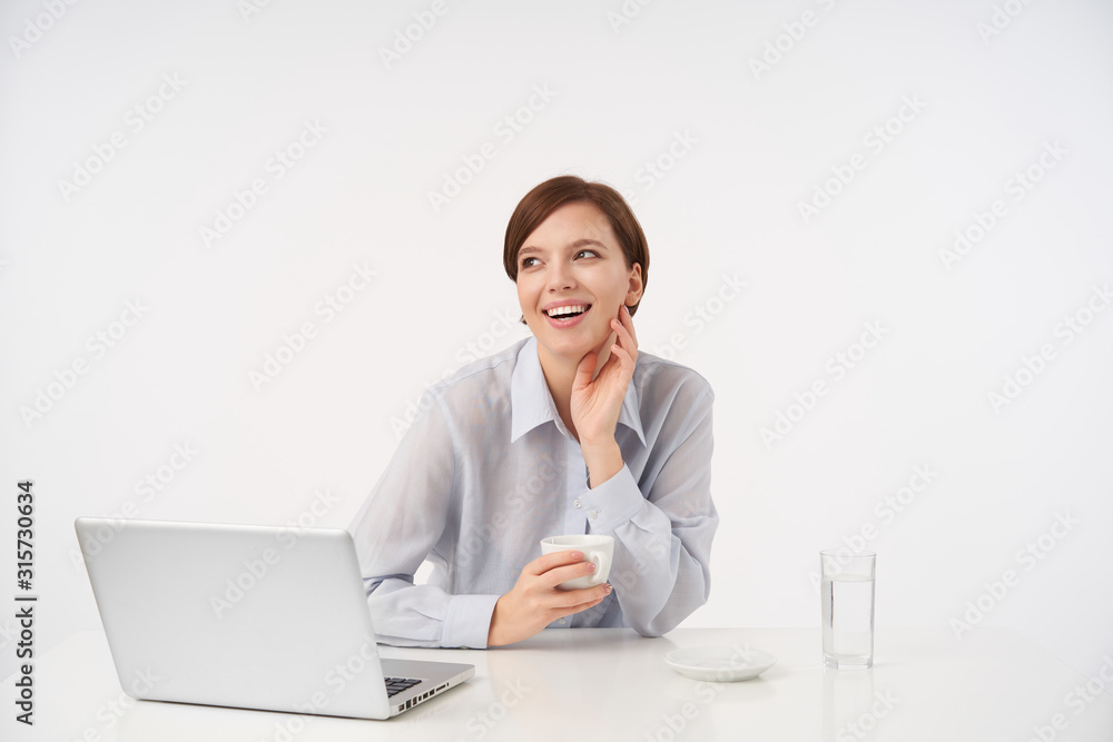 Good looking young cheerful brown haired woman with short trendy haircut touching her neck with raised hand and smiling happilly, posing over white background with cup of tea Good looking young cheerful brown haired woman with short trendy haircut touching her neck with raised hand and smiling happilly, posing over white background with cup of tea